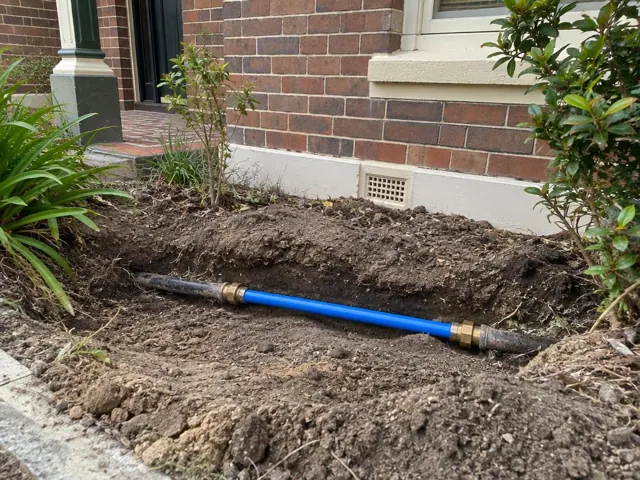 Exposed repaired water pipe in a dug trench beside a brick house wall in Adamstown